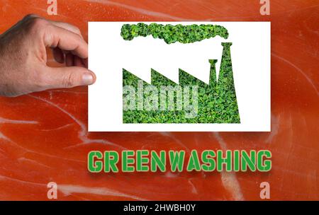 Greenwashing: A man`s hand holds a white sheet of paper with the outline of a factory cut out with green grass inside.Background with ham meat Stock Photo